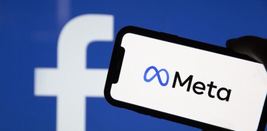 Meta (dawny Facebook) - zbliżenie na ekran smartfona. Na białym tle duże logo Mety. N drugim planie niebieska ściana z białym logo facebooka.