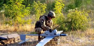 bezzałogowce flyeye - na leśnej polanie, ubrany w maskujący strój żołnierz przygotowuje do startu drona FlyEye. Dron wygląda jak duży zabawkowy model samolotu.