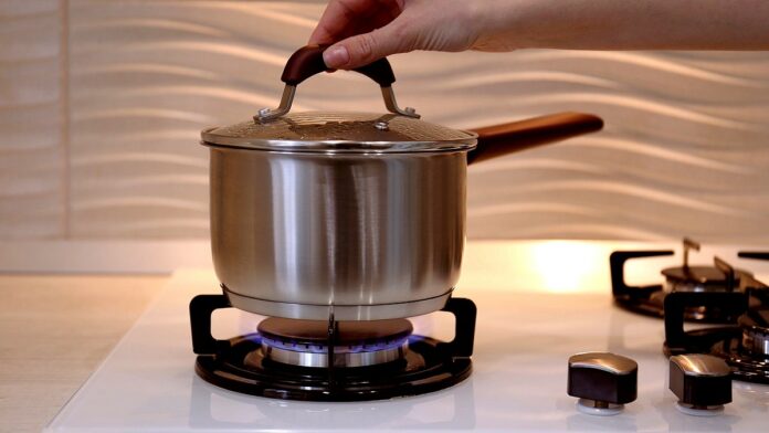 ceny gazu - zbliżen9ie na kuchenkę gazową. Na płonącym palniku stoi srebrny garnek z pokrywą. Widać rękę trzymającą pokrywę.