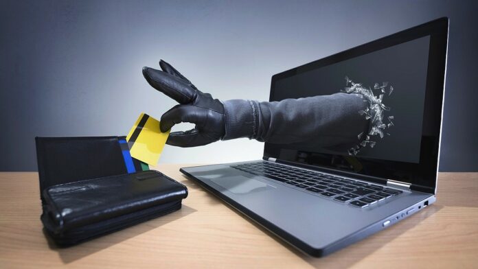 numer PESEL - z ekranu laptopa wysuwa się ręka w czarnej rękawiczce i kradnie kartę kredytową z leżącego obok portfela.