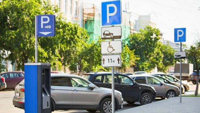 roztargnienie kierowcy z Warszawy - widok słonecznej ulicy i zaparkowanych na niej aut. Na pierwszym planie widać niebieski parkometr, a dalej rząd samochodów oraz kilka znaków informujących o miejscu do parkowania.