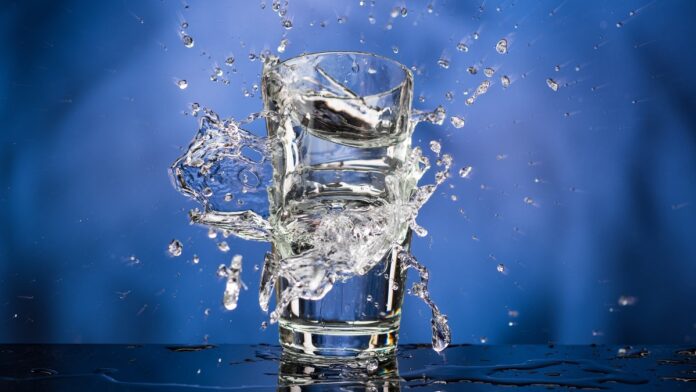 wyższe ceny wody - na niebieskim tle stoi szklanka, do której z dużej wysokości wlewana jest woda. Tworzy malowniczy rozbryzg.